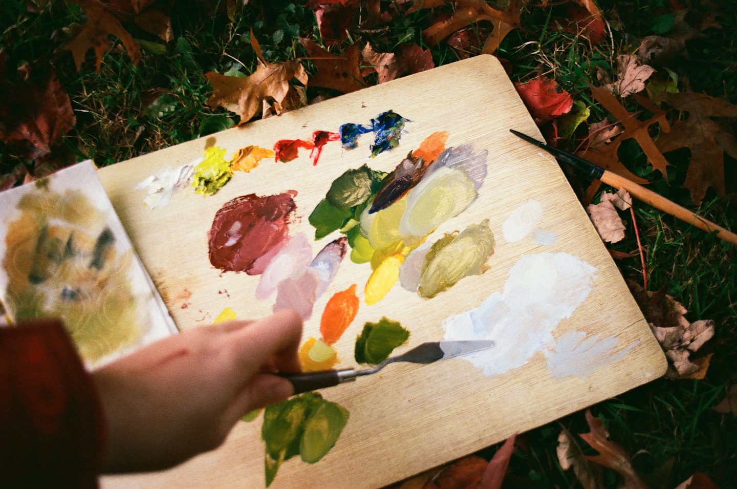Eine Hand mischt bunte Farben auf einer Holzpalette im Freien.